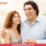 A smiling woman with curly red hair and a tall man with curly dark hair embracing, set against tropical foliage.