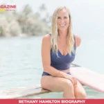 Bethany Hamilton, a surfer with one arm, sitting on a wooden dock beside a pink surfboard