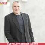 Rudy Ruettiger net worth article image of a smiling Rudy Ruettiger in a black blazer holding a camera