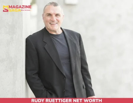Rudy Ruettiger net worth article image of a smiling Rudy Ruettiger in a black blazer holding a camera