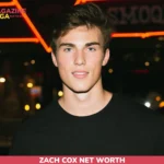 Zach Cox in a black t-shirt at night in front of a red neon sign for the net worth article
