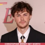 Christopher Briney in a black suit with a gold chain-link tie clasp at an event backdrop