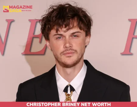 Christopher Briney in a black suit with a gold chain-link tie clasp at an event backdrop