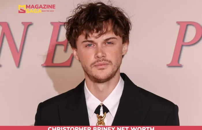 Christopher Briney in a black suit with a gold chain-link tie clasp at an event backdrop