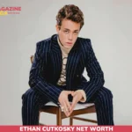 Ethan Cutkosky net worth article portrait, seated in a navy pinstripe suit on a wooden chair