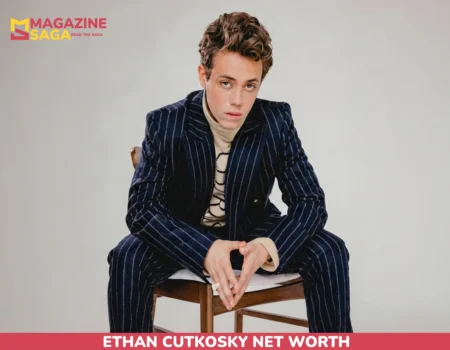 Ethan Cutkosky net worth article portrait, seated in a navy pinstripe suit on a wooden chair